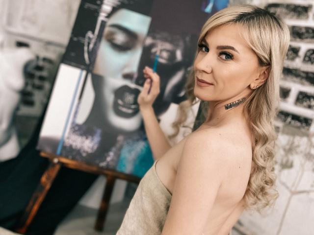 ParadiseRoxanne with blonde curls, looking over her shoulder, holding a paintbrush against black-and-white portraits, wearing a strapless beige top, showing her tits, smiling subtly.
