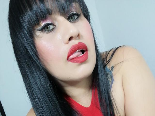 CathyCat a les cheveux noirs, du rouge à lèvres et un fard à paupières rose brillant. Elle sort sa langue, qui est légèrement visible. Son haut rouge révèle un tatouage sur son épaule..