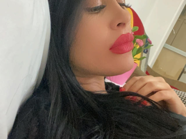 Azazelle's face with dark black hair, red lipstick, and closed eyes, lying down, right hand touching her neck. She's wearing a black top with red accents.