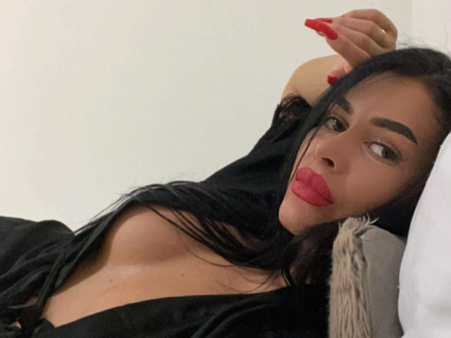 Azazelle with long black hair, red lips, and red nails, lying on a bed, wearing a black top that reveals her cleavage, looking at the camera.