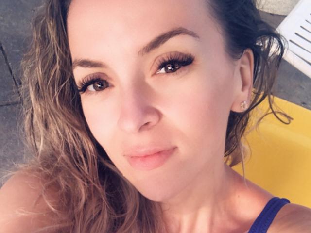 SophieDeee has wavy brown hair, thick eyelashes, and a slight smile. She's wearing a purple top, showing a bit of her shoulder, with small earrings.