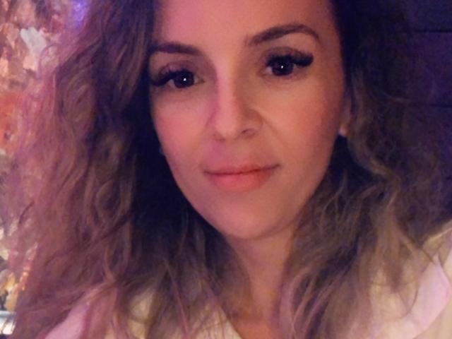 SophieDeee has curly brown hair, subtle makeup with long eyelashes, and a slight smile. Her pinkish skin and small nose are visible. She's wearing a white top.