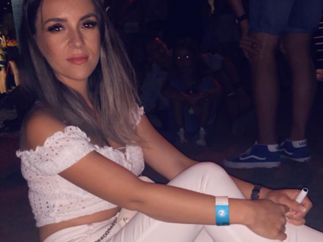 SophieDeee with gray hair, white off-shoulder top, white pants, and blue watch, holding a cigarette, sitting with legs crossed, looking at camera, wearing subtle makeup.