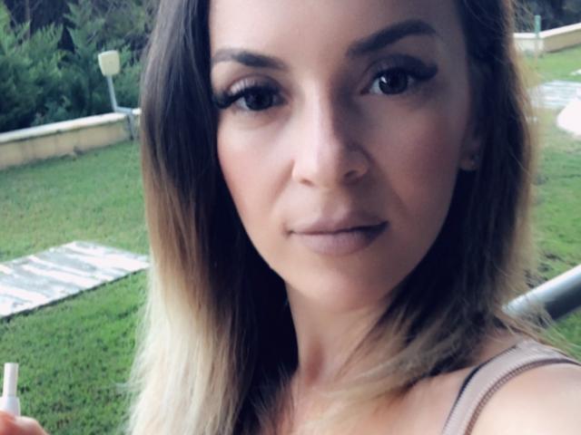 SophieDeee has light brown hair, subtle makeup, and is looking at the camera with a slight smile, holding a straw in a grassy outdoor setting.
