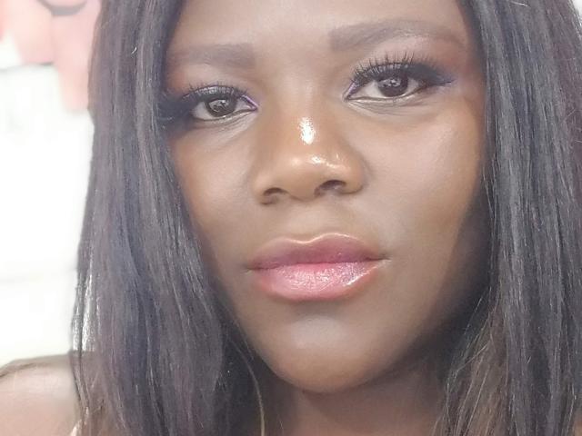 Le visage éclatant de NalaBacker, avec sa peau brun foncé, ses longs cheveux noirs, son fard à paupières violet, ses cils épais et ses lèvres roses brillantes. Son expression est neutre, et elle regarde directement l'appareil photo..