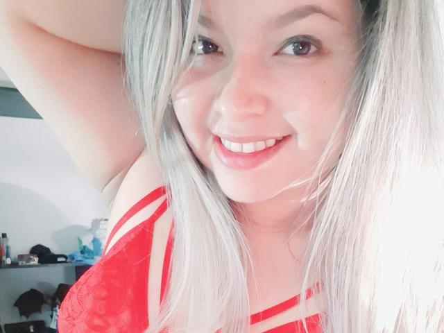Myalewwis, con i capelli argentati, sorridente, indossa lingerie di pizzo rossa, con il seno visibile, braccio alzato, guarda la fotocamera, sfondo bianco..