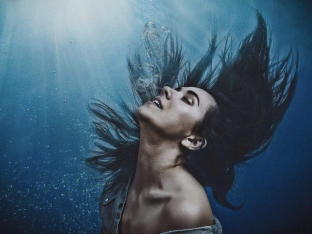 Misstyk with long dark hair flowing upward, eyes closed, and mouth slightly open, underwater with light beams and bubbles. She's topless, showing her bare shoulders and collarbone.