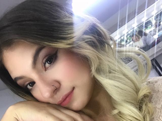 Valerie Martens, con cabello rubio y ondulado, un piercing en la nariz y un maquillaje discreto, apoya su barbilla en la mano y sonríe suavemente. Sus senos son ligeramente visibles, pero en su mayoría están cubiertos..