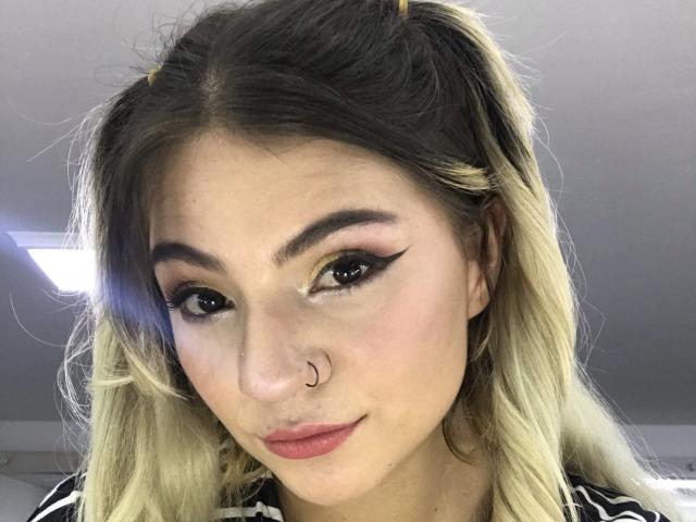 ValerieMartens ha i capelli biondi con sfumature, un trucco occhi nero con eyeliner allungato, rossetto rosa, un piercing al naso e un sorriso carino e sicuro. Indossa una camicia a righe con maniche a sbuffo..