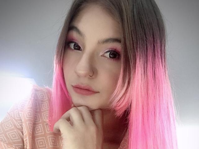 ValerieMartens tem cabelo com degradê rosa, sombra rosa nos olhos, um piercing no nariz e está olhando para a câmera com a mão embaixo do queixo, vestindo uma blusa rosa com estampa..