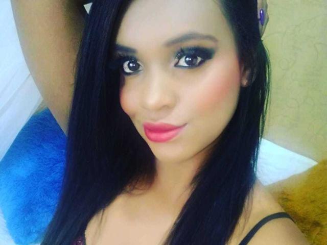 AllyCharlotee has long black hair, dark eyeliner, pink lipstick, and a cute smile. She's wearing a black strap top, showing her small tits. Her skin is tan.