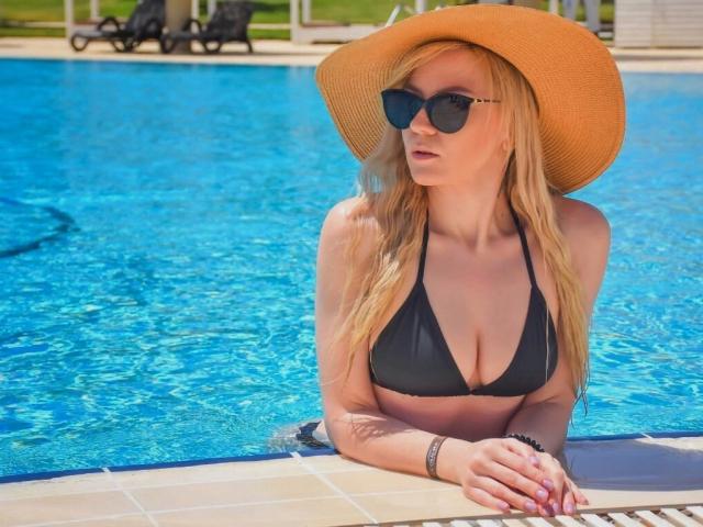 ParadiseRoxanne with blonde hair, wearing a black bikini top, sunhat, and sunglasses, leaning on a pool edge, showing her tits and nipples, with blue water behind her.