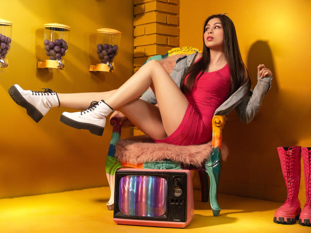 IsaTimeSweet met lang bruin haar, een roze jurk, witte laarzen en roze laarzen, met de benen omhoog, zittend op een kleurrijke stoel, naast een vintage televisie, een gele muur en paarse druiven..