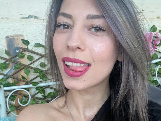 PoxyVibe's face with pink lipstick, tongue out, brown eyes, long eyelashes, straight brown hair, fair skin, and a small mole under her right eye. She's smiling at the camera.