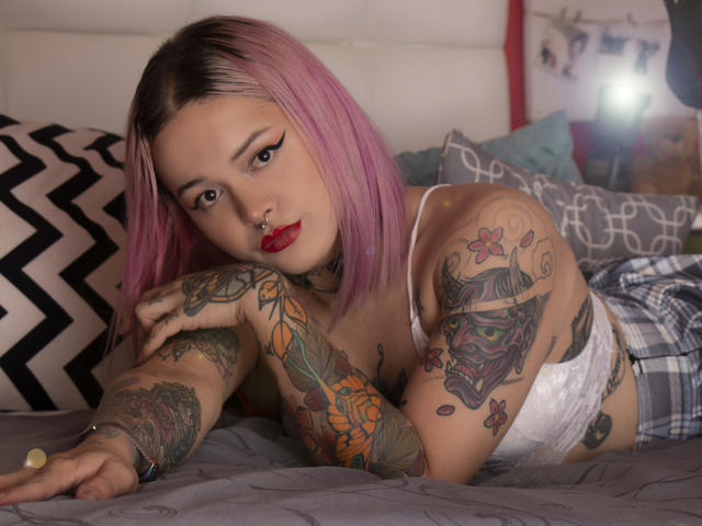 NiNNastormx met roze haar, rode lippenstift en tatoeages, liggend op een bed in witte kant lingerie, kijkend naar de camera met een neusring, met getatoeëerde armen die rusten..