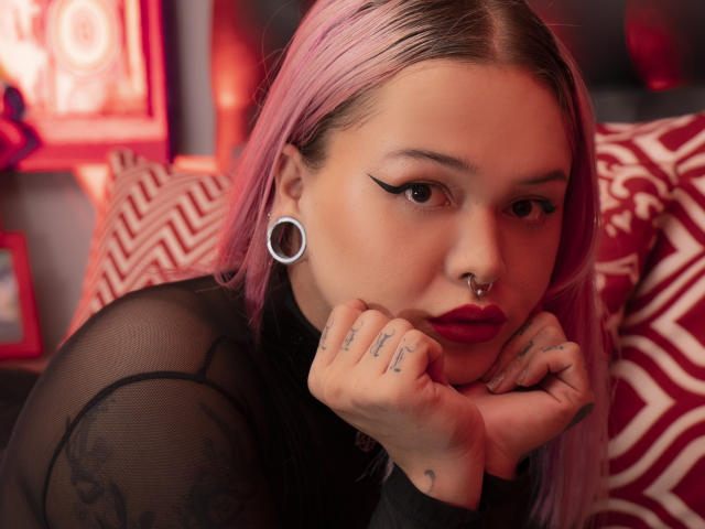 NiNNastormx a les cheveux roses, un trait d'eye-liner noir, du rouge à lèvres rouge, un anneau au nez, des élargisseurs d'oreilles, un haut noir transparent, des tatouages sur les mains et un coussin rouge à motifs en arrière-plan..