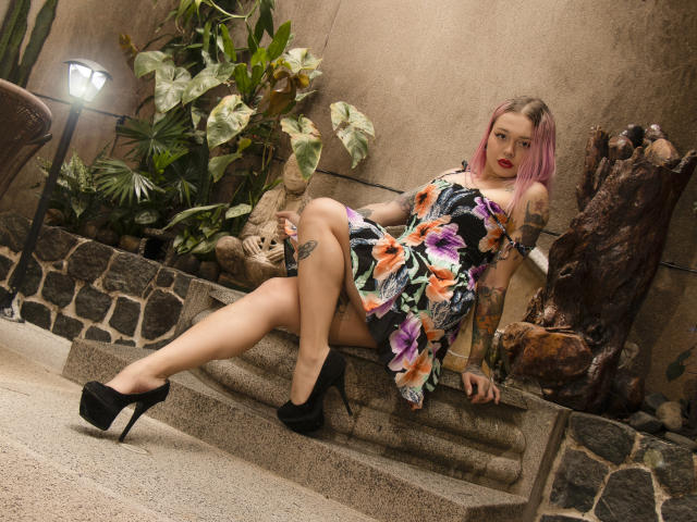 NiNNastormx met roze haar, tatoeages, een bloemenjurk en zwarte hakken, zittend op stenen treden, benen gespreid, met een tatoeage van een geslachtsdeel zichtbaar, rode lippenstift en een zelfverzekerde blik..