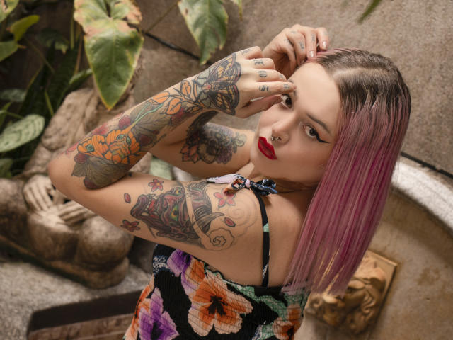 NiNNastormx with pink hair, red lipstick, and tattoos on arms and chest, wearing a colorful floral dress, touching her face, showing off her tattoos and colorful nails.