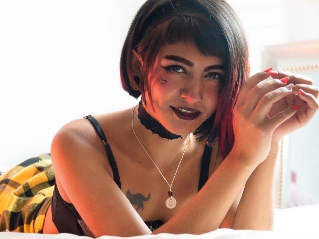 SerenaDeStark com cabelo curto preto e vermelho, colar preto, batom escuro, sutiã preto, tatuagem abaixo do seio, unhas vermelhas, deitada na cama, cobertor xadrez, sorrindo, segurando as mãos, colar..