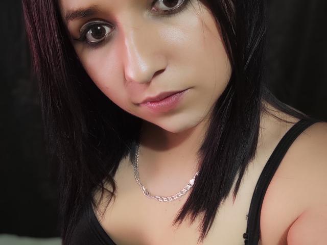 Kkatalina, com olhos castanho-escuros, pele clara e cabelo preto e liso, vestindo uma regata preta e um colar de prata, olhando ligeiramente para baixo com um sorriso discreto..