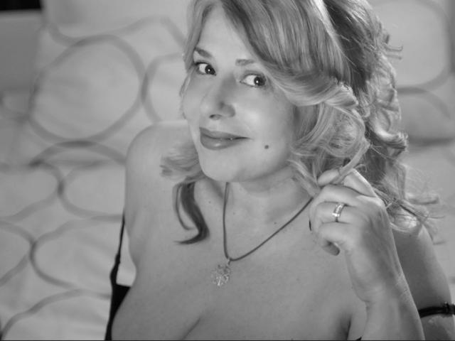Black-and-white photo of ByaStorm with curly blonde hair, smiling, holding her hair, showing her cleavage, wearing a necklace, ring, and black strap, looking up.