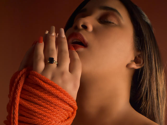 Il viso di SamantaSub, con gli occhi chiusi e la bocca leggermente aperta, una mano con una corda arancione, un anello nero e unghie arancioni che toccano le sue labbra, sfondo rosso, capelli castano scuro, luce soffusa, espressione sensuale..