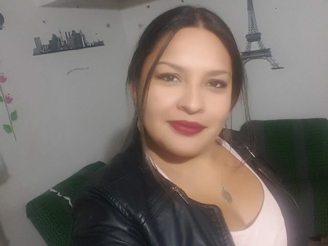 Adi69, con pechos de tamaño medio, vestida con una chaqueta de cuero negra y una camiseta rosa, tiene el cabello oscuro recogido en una coleta, lápiz labial rojo y un collar de plata, y está sonriendo en una foto..