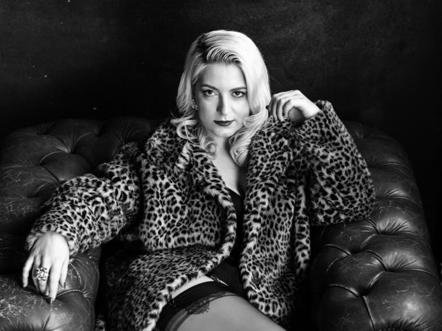 FeetSeductress hat blondes Haar, einen Leopardenfellmantel und dunkle Dessous, und liegt auf einem gepolsterten Ledersofa, mit einem verführerischen Ausdruck, wobei sie ihren Ring und ihren Oberschenkel zeigt..