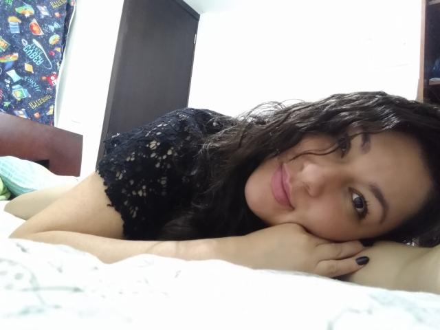 SakotoDika with curly black hair, wearing a black lace top, lying on a bed, head on hands, smiling at the camera, showing her cute face and medium tits.