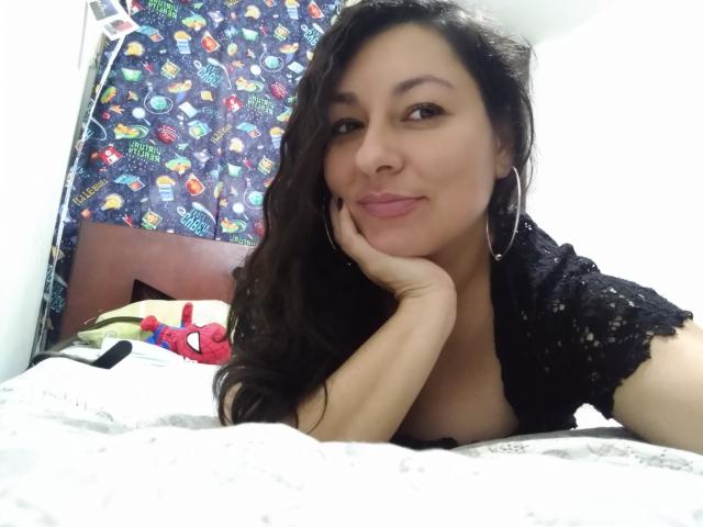 SakotoDika, met lang zwart haar, ligt op een bed, haar hoofd rustend op haar hand, gekleed in een zwarte kanten top, met oorringen en een subtiele glimlach, waarbij haar borsten zichtbaar zijn..