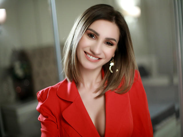 JuliaRichard with shoulder-length brown hair, smiling, wearing a red blazer with a low neckline, showing a hint of her tits, and gold earrings, in a bright room.