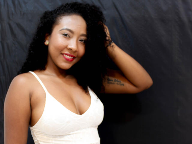 CaroldelaTorre, con i capelli neri ricci, sorridente, indossa un top bianco in pizzo che mette in mostra il seno, rossetto rosso, un piccolo tatuaggio sul braccio destro, sfondo nero..
