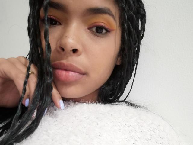 VickyGreys has light brown skin, black braids, orange-yellow eyeshadow, purple eyeliner, pink lips, light purple nails, white fuzzy top, and a gold ring on her left hand.