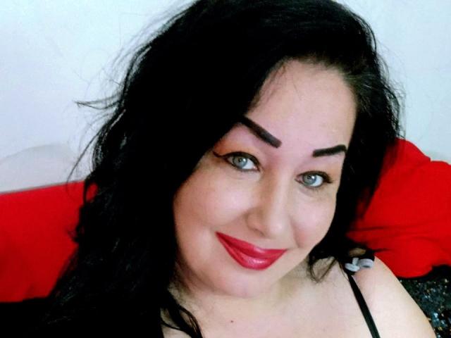 LexyRose has black hair, thick eyebrows, blue eyes, and red lipstick. She's smiling with a cute face, wearing a black top. Her tits are not visible.