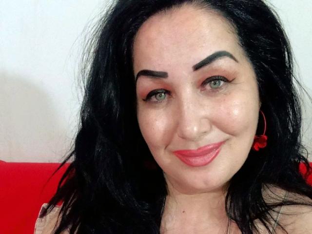 LexyRose has black hair, green eyes, and red lipstick, with thick black eyebrows, wearing red earrings, smiling with a slightly flushed face. She's a cute chick.