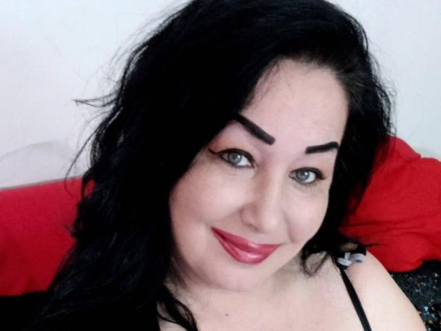 LexyRose met zwart haar, blauwe ogen, rode lippenstift en zwarte wenkbrauwen, lachend naar de camera. Ze draagt een zwarte top, en haar borsten zijn gedeeltelijk zichtbaar..