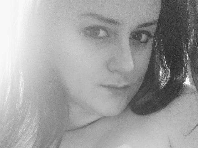 Selfie en blanco y negro de CatrinBlackFox con cabello largo y oscuro, un piercing en la nariz y una sonrisa sutil, mostrando sus hombros desnudos y un collar delgado. Sus ojos están ligeramente entrecerrados..