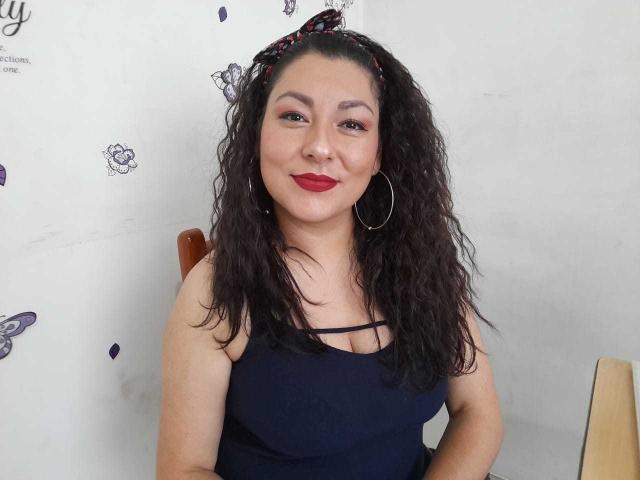 SakotoDika has curly black hair, red lipstick, hoop earrings, and a dark blue tank top showing her tits. She's smiling, looking at the camera.