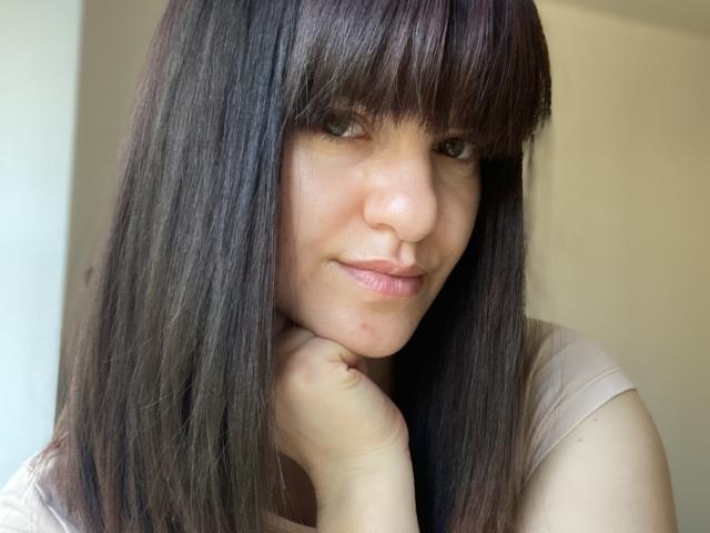 LaraFontaineX has straight brown hair with bangs, a light skin tone, and a subtle smile, resting her chin on her hand, wearing a beige shirt.