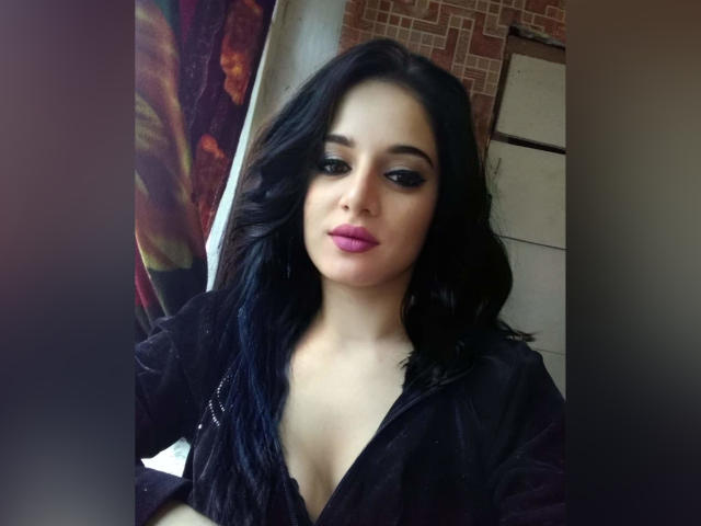 KateLaAngel tiene el cabello largo y negro, lápiz labial rosa y maquillaje de ojos oscuro. Lleva una camisa negra que deja ver parte de su escote. Su expresión es ligeramente seria, mirando a la cámara..