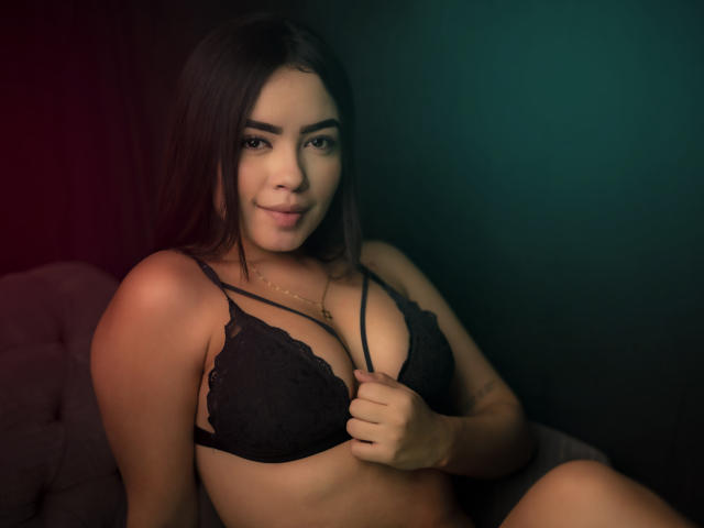 AlanaPierce, with long dark hair, small nose ring, and slight smile, wears a black lace bra, holding it, showing her tits, in a dimly lit, green-to-red gradient room.
