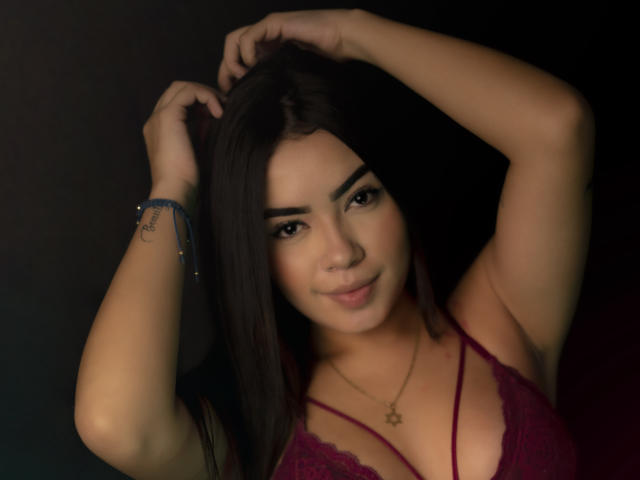 AlanaPierce with dark hair, small tits in a red lace bra, arms raised, smiling, wearing a necklace, bracelet with a tattoo on her wrist. Soft lighting, dark background.