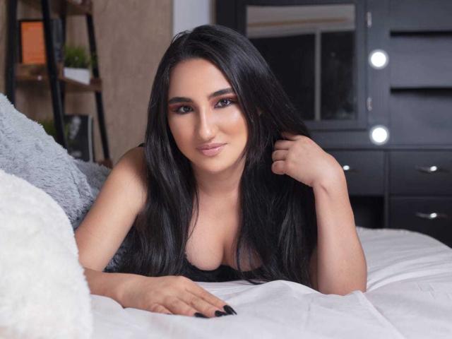 Zara Moretti, com longos cabelos pretos, deitada em uma cama, mostrando os seios, vestindo uma blusa preta, olhando para a câmera com um leve sorriso, com a mão no cabelo..