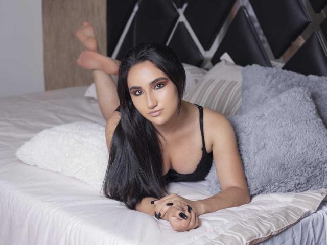 Zara Moretti, com longos cabelos pretos, está deitada em uma cama, vestindo uma regata preta que revela seus seios, e olhando para a câmera com um leve sorriso. Suas pernas estão levantadas..