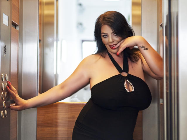 YulliaMilfX with big tits, black halter dress, cutout, red lipstick, black hair, right hand on door, left hand touching chin, wooden elevator, tattoo on right wrist.