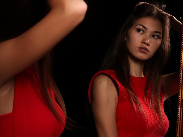 KiraFendy con lunghi capelli castani, vestito a coste rosso, espressione seria, che alza un braccio di fronte a uno specchio, riflettendo la sua immagine, sfondo nero, seno visibile, attraente, magra, sexy, trucco leggero..