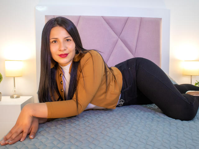 Bethany Roberts, met lang zwart haar, een bruin shirt, zwarte jeans en rode lipstick, ligt op een blauw geborduurd bed, lachend naar de camera, waarbij haar schattige billen en zachte borsten zichtbaar zijn..