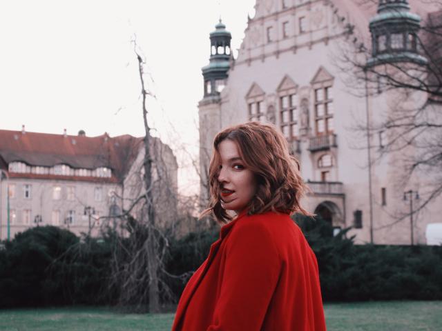 KenzieONEx com cabelo castanho ondulado, casaco vermelho, grama verde e um edifício histórico ao fundo, olhando por cima do ombro com um leve sorriso..