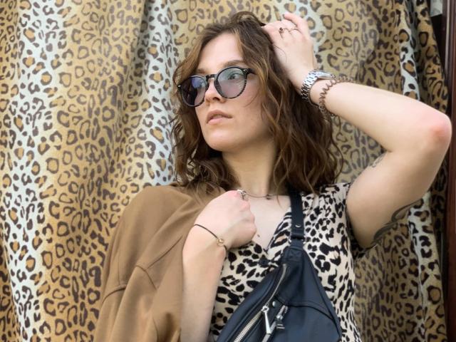 KenzieONEx heeft golvend bruin haar, draagt een donker paar zonnebril, een top met luipaardprint, en een bruine jas, met een zwarte tas, een zilveren horloge en een armband..