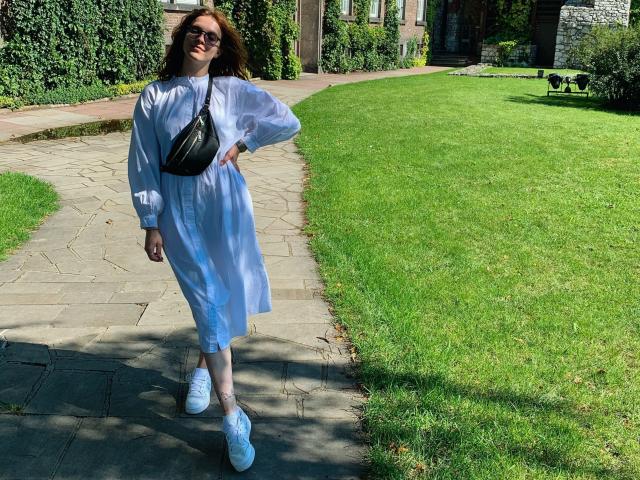 KenzieONEx con cabello rojo, gafas, vestido azul, bolso bandolera negro, zapatillas blancas, de pie en un camino de piedra, césped verde y un edificio de ladrillo en el fondo, sonriendo..